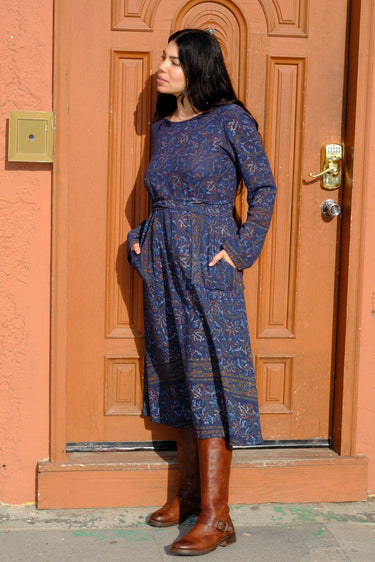 A woman standing in front of a door, wearing a blue tapestry wrap dress with long bell sleeves and a quarter circle skirt, featuring patch pockets.