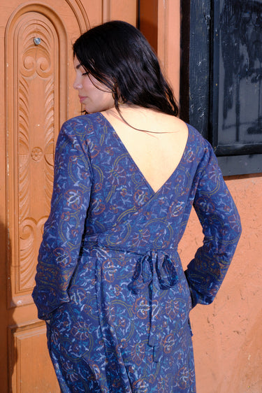 A woman standing in front of a door, wearing a blue tapestry wrap dress with long bell sleeves and a quarter circle skirt, featuring patch pockets. Back View.