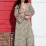 A woman standing in front of a red wall, wearing a green and blue floral maxi dress with a zip front and long full sleeves.