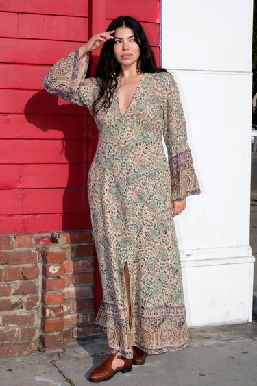 A woman standing in front of a red wall, wearing a green and blue floral maxi dress with a zip front and long full sleeves.
