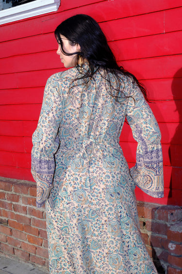 A woman standing in front of a red wall, wearing a green and blue floral maxi dress with a zip front and long full sleeves, back view.