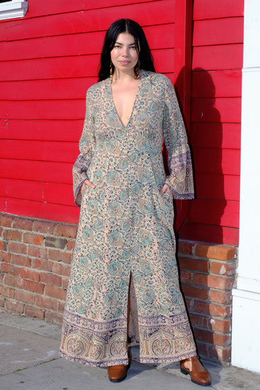 A woman standing in front of a red wall, wearing a green and blue floral maxi dress with a zip front and long full sleeves.