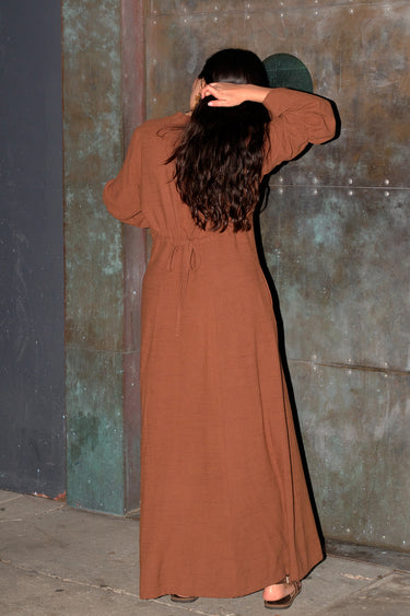 A woman standing by a metal door wearing a nutmeg brown zip front long sleeve dress, back view.