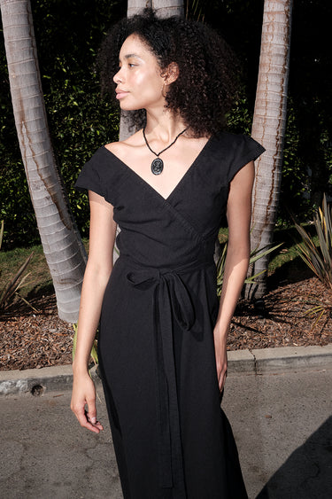 A woman standing outside wearing a long black maxi wrap dress with an empire waist and an elastic neckline.