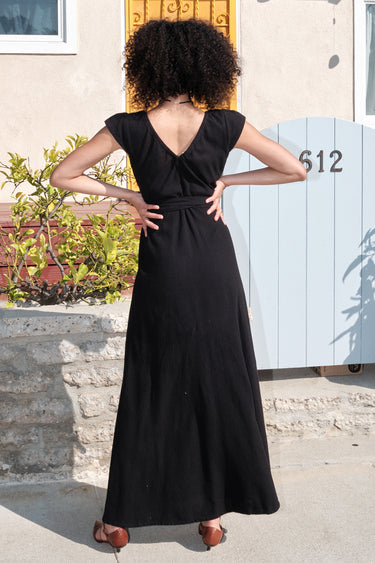 A woman standing outside wearing a long black maxi wrap dress with an empire waist and an elastic neckline. Cack view.