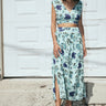 A woman standing outside by a white garage wearing an aqua ground floral tapestry skirt with a drop waist and side slits, paired with a matching cocoa top. 