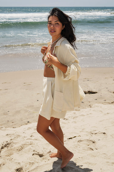 A woman standing on a beach wearing a floral tapestry bikini top and an alabaster trouser short, with a matching alabaster shirt jacket. Side View