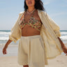 A woman standing on a beach wearing a floral tapestry bikini top and an alabaster trouser short, with a matching alabaster shirt jacket.