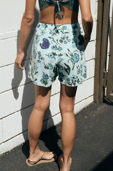 A woman standing outside wearing an aqua ground floral tapestry short with dark blue paisley print bra top. Back View