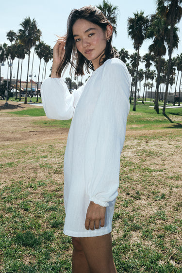 A woman wearing a white mini dress with long sleeves and a gathered neckline, standing in an outdoor setting with palm trees in the background. Side View