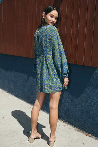 a woman standing outside wearing a dark blue paisley tapestry mini dress with long sleeves. Back View