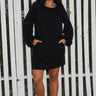 A woman standing in front of a white fence, wearing a black mini dress with a gathered neckline, long full sleeves, and a split back. The dress is made of a crinkle textured cotton fabric.