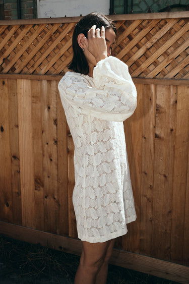 A woman standing outside by a brown wood fence wearing a lace mini dress, side view. 