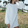 A woman wearing a white mini dress with long sleeves and a gathered neckline, standing in an outdoor setting with palm trees in the background.