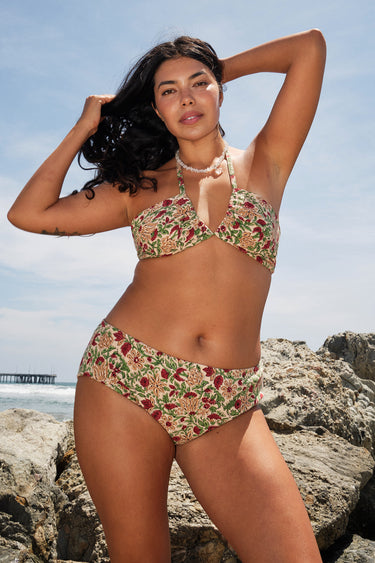 A woman on a beach by rocks wearing a beige ground with red, brown and green floral tapestry bikini bottom with a high rise and matching bikini top.