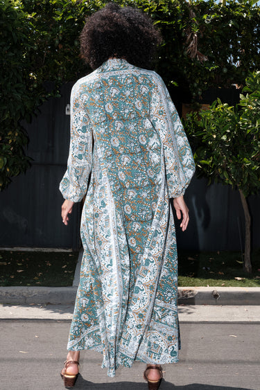 Woman wearing a long tarragon colored floral tapestry caftan standing outdoors with greenery in the background. Back View
