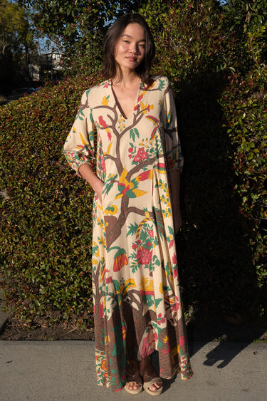 A woman standing outside by greenery wearing a maxi caftan cut in a tree of life handloom tapestry.