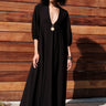 A woman standing outside by a brown wood fence wearing a maxi length black v neck caftan.