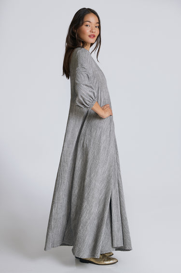A woman standing in a white walled photo studio wearing a 3/4 sleeve maxi caftan cut from a salt and pepper textured cotton, side view.