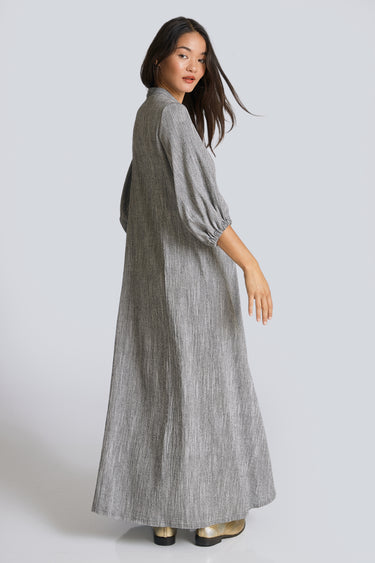 A woman standing in a white walled photo studio wearing a 3/4 sleeve maxi caftan cut from a salt and pepper textured cotton, back view.