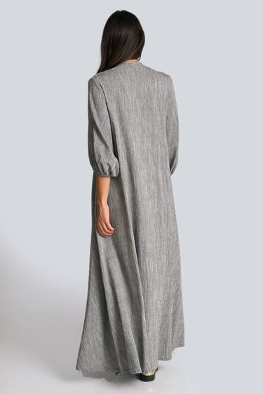 A woman standing in a white walled photo studio wearing a 3/4 sleeve maxi caftan cut from a salt and pepper textured cotton, back vew.