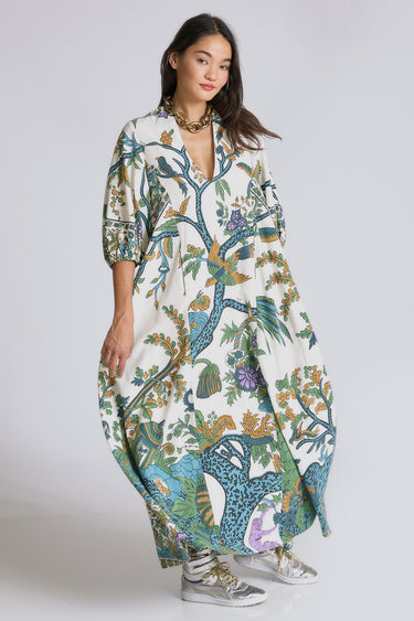 A model standing in a white photo studio wearing a maxi caftan, with three quarter balloon sleeves and  pockets. The caftan is cut from a blue and green hand block printed tree of life tapestry