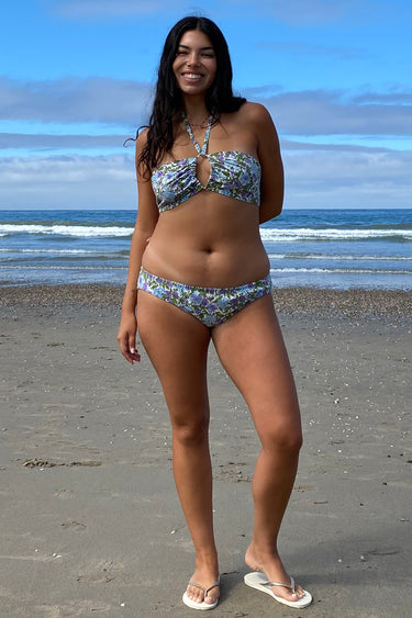 A woman on a beach wearing a white ground with lavender and blue floral tapestry bikini bottom with a low-rise design and matching bikini top.
