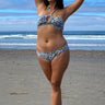 A woman on a beach wearing a white ground with lavender and blue floral tapestry bikini bottom with a low-rise design and matching bikini top.