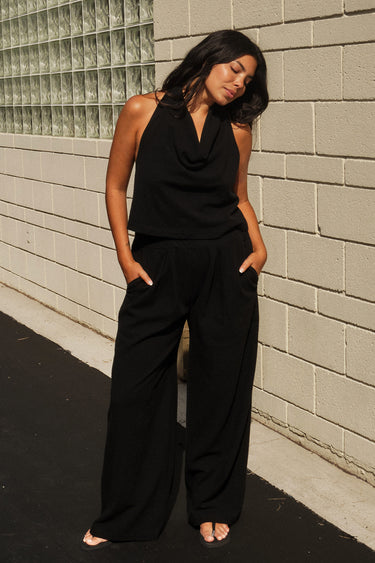 A woman standing  outside next to an off white building wearing black trousers and a matching halter top.