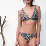 A woman standing outside by a white wall wearing a stripe blue and pink floral floral handloom tapestry bikini bra top with matching low rise bikini bottoms