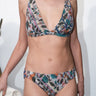 A woman standing outside by a white wall wearing a stripe floral handloom tapestry bikini with low rise bottom with a matching bra top. 