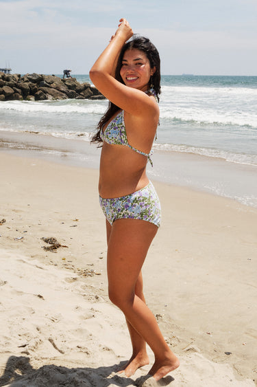 A woman on a beach wearing a white ground with lavender, blue and green floral tapestry bikini bottom with a high rise and matching bikini top. side view