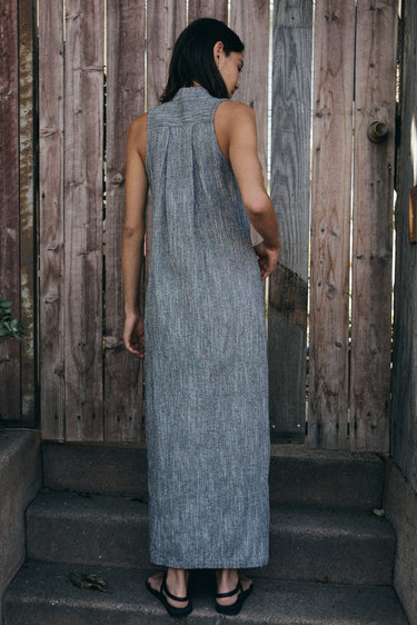 A model wearing a gray maxi shift dress with a front zipper and pockets, standing near a wooden fence. Back View