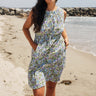 A woman standing on a beach wearing a knee-length floral dress with broomstick pleats, featuring an elastic neckline and a slit back. The dress is a white ground tapestry with blue and lavenders flowers.