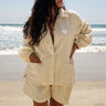 A woman standing on a beach wearing an alabaster unlined shirt jacket with big patch pockets and an elastic shirred sleeve cuff, with matching shorts.