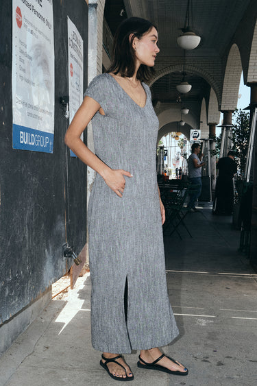 Woman standing on the sidewalk, outside wearing a salt and pepper yarn dye long sheath dress with a cap sleeve.  Side View.