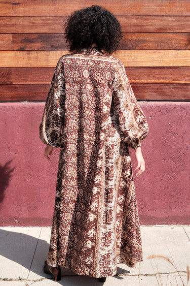 A woman standing by a red wall, outside wearing a long sleeve maxi caftan cut in a raisin floral tapestry, back view.