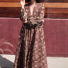 A woman standing by a red wall, outside wearing a long sleeve maxi caftan cut in a raisin floral tapestry.