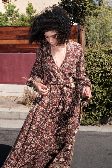 A woman standing by a red wall, outside wearing a long sleeve maxi caftan cut in a raisin floral tapestry, belted.