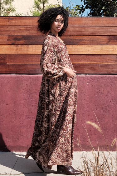 A woman standing by a red wall, outside wearing a long sleeve maxi caftan cut in a raisin floral tapestry, side view. 