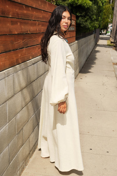 Woman standing by an on sidewalk wearing a long sleeve alabaster maxi wrap dress, side view. With a turquoise necklace.