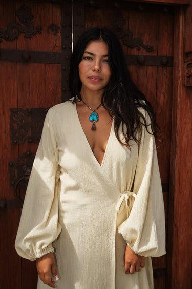 Woman standing by an ornate gate wearing a long sleeve alabaster maxi wrap dress. With a turquoise necklace.