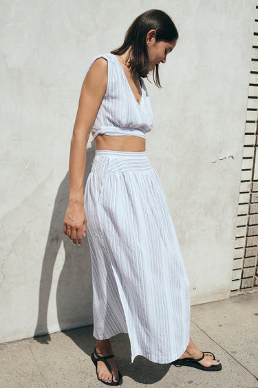 A woman standing outside wearing a white gauze stripe skirt with side slits and a drop waist, paired with a matching cocoa-colored top. 