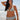 A woman on a beach wearing a white ground with lavender, blue and green floral tapestry bikini bottom with a high rise and matching bikini top.