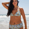 A woman on a beach wearing a white ground with lavender, blue and green floral tapestry bikini bottom with a high rise and matching bikini top.