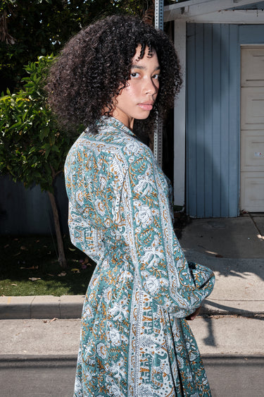 Woman wearing a long tarragon colored floral tapestry caftan standing outdoors with greenery in the background. Belted back view.