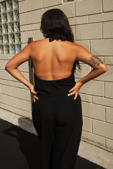 A woman standing outside next to an off white building wearing black trousers and a matching halter top. Back View