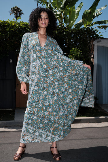 Woman wearing a long tarragon colored floral tapestry caftan standing outdoors with greenery in the background.
