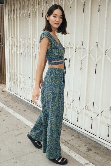 A woman wearing a blue paisley tapestry trouser with a matching crop top, standing outside by a white security gate, side view. 