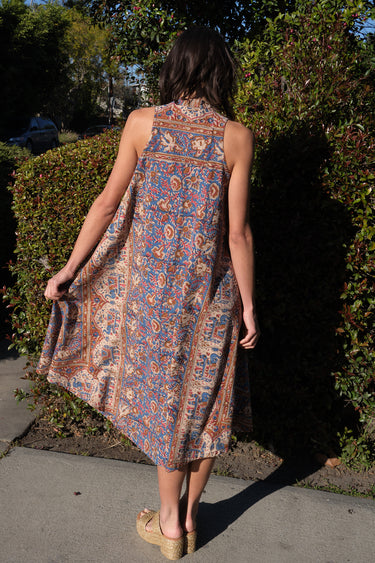 A woman standing in front of greenery outside wearing a sleeves midi caftan cut in a blue floral tapestry. Back View. 